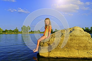 Cute little girl in a bathing suit sitting on a large rock by the lake at sunset. Summer and happy childhood concept