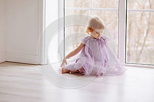 A cute little girl in a beautiful dress sits by the window in a bright sunny room