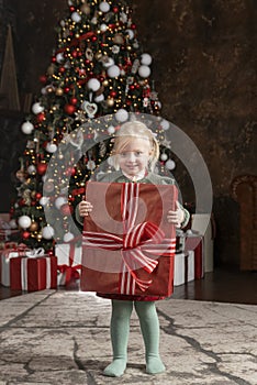 Cute little girl with big red gift box. Smiling blonde child near Christmas tree. Christmas Eve