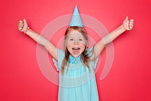 Cute little girl with Birthday hat showing thumb-up gesture on color background