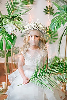 Cute little girl with blond curly hair in a white wedding dress and a wreath of flowers in floral decorations