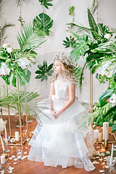 Cute little girl with blond curly hair in a white wedding dress and a wreath of flowers in floral decorations