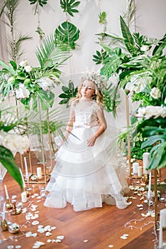 Cute little girl with blond curly hair in a white wedding dress and a wreath of flowers in floral decorations