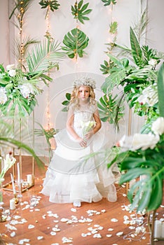 Cute little girl with blond curly hair in a white wedding dress and a wreath of flowers in floral decorations