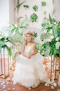 Cute little girl with blond curly hair in a white wedding dress and a wreath of flowers in floral decorations