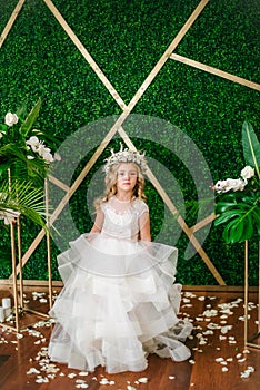 Cute little girl with blond curly hair in a white wedding dress and a wreath of flowers in floral decorations