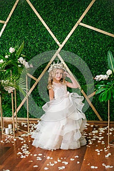 Cute little girl with blond curly hair in a white wedding dress and a wreath of flowers in floral decorations