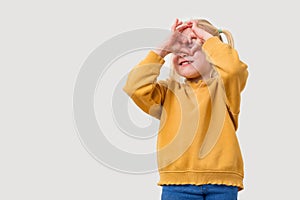 A cute little girl with blond hair in a yellow sweater closes her eyes with her hands and makes a heart gesture. Isolated on white