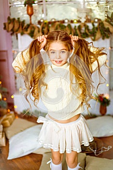 Cute little girl with blond long curly hair in a light knitted sweater by the festive fireplace with garlands