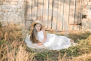 Cute little girl with blond long hair in a summer field at sunset with a white dress with a straw hat