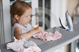 Cute little girl with blonde hair is leaning on ironing clothes on board at home. Daughter helping to mother ironing clothes for