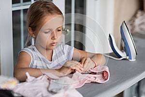 Cute little girl with blonde hair is leaning on ironing clothes on board at home. Daughter helping to mother ironing clothes for