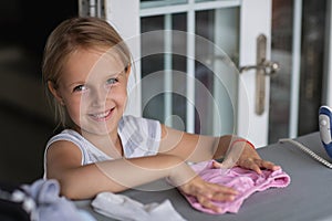 Cute little girl with blonde hair is leaning on ironing clothes on board at home. Daughter helping to mother ironing clothes for