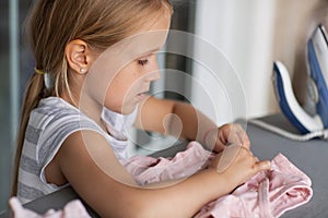 Cute little girl with blonde hair is leaning on ironing clothes on board at home. Daughter helping to mother ironing clothes for