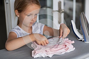 Cute little girl with blonde hair is leaning on ironing clothes on board at home. Daughter helping to mother ironing clothes for