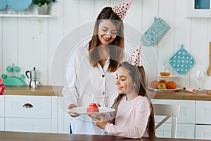 Cute little girl blowing out candles on birthday cake on kitchen with mother. Celebrating birthday at home.