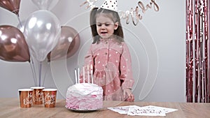 Cute little girl blows out candles on a birthday cake at home against a backdrop of balloons. Child's birthday.