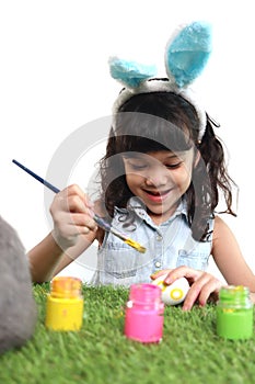 Cute little girl with blue bunny ears painting eggs with brush on green grass meadow, joyful kid celebrating Easter holiday on