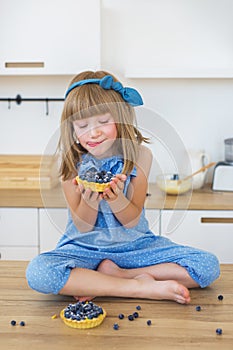 Cute little girl in blue dress sits on a table with a cake and looks on it and licks her lips