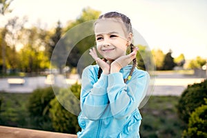 A cute little girl in a blue jacket with pigtails poses while sitting on a bench with her hands pressed to her face