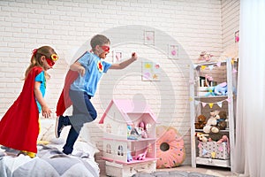 Cute little girl and boy jumping from bed to fly, play superhero with cloak and mask at home in kids bedroom