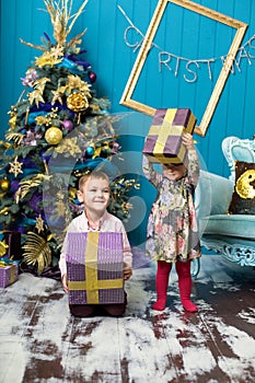 Cute little girl and boy are smiling and holding gifts under the Christmas tree. Brother and sister unpack gift boxes on Christmas