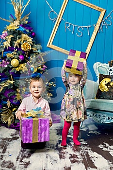 Cute little girl and boy are smiling and holding gifts under the Christmas tree. Brother and sister unpack gift boxes on Christmas