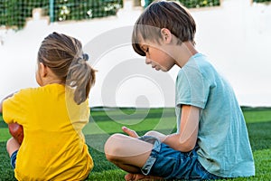 Cute little girl with brother sitting together thoughtfully on green grass and looking away