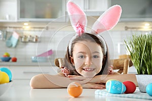 Cute little girl with bunny ears headband and painted Easter eggs sitting at table in kitchen