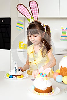 A cute little girl with bunny ears prepares an Easter cake and painted eggs. Religious holiday