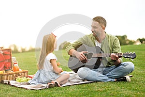 Cute little girl in casual clothes sitting on a green grass in park and looking at her loving dad playing guitar