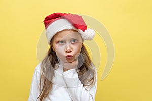 Cute little girl in a Christmas hat. The girl shows emotions on a yellow background. New year concept