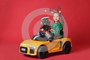 Cute little girl with Christmas tree driving children`s electric toy car on red background