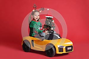 Cute little girl with Christmas tree driving children`s electric toy car on red background