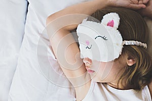 Cute little girl closeup sleeping in bed wearing white cat plush sleep mask. Early morning wake up, rise to kindergarten, school.