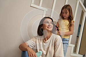 Cute little girl combing long hair of her beautiful smiling mother, mom and daughter spending time together at home