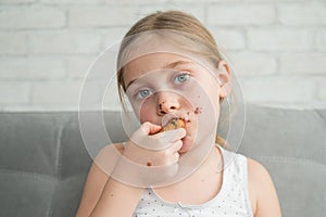 A cute little girl covered in chocolate eats cookies while sitting on the sofa.