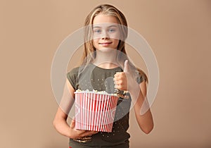 Cute little girl with cup of popcorn showing thumb-up on color background