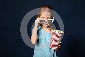 Cute little girl with cup of popcorn wearing 3D cinema glasses on dark background