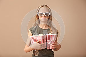 Cute little girl with cups of popcorn wearing 3D cinema glasses on color background
