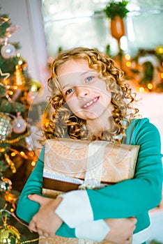 Cute little girl with curly blond hair at home near a Christmas tree with gifts and garlands and a decorated fireplace