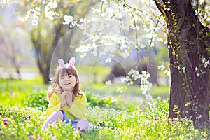 Cute little girl with curly hair wearing bunny ears and summer dress having fun during Easter egg hunt relaxing in the garden