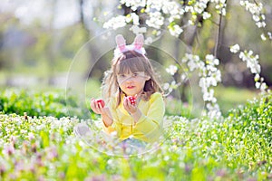 Cute little girl with curly hair wearing bunny ears and summer dress having fun during Easter egg hunt relaxing in the garden