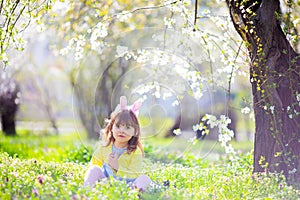 Cute little girl with curly hair wearing bunny ears and summer dress having fun during Easter egg hunt relaxing in the garden