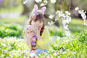 Cute little girl with curly hair wearing bunny ears and summer dress having fun during Easter egg hunt relaxing in the garden