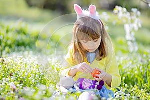 Cute little girl with curly hair wearing bunny ears and summer dress having fun during Easter egg hunt relaxing in the garden