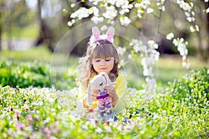 Cute little girl with curly hair wearing bunny ears and summer dress having fun during Easter egg hunt relaxing in the garden