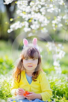 Cute little girl with curly hair wearing bunny ears and summer dress having fun during Easter egg hunt relaxing in the garden