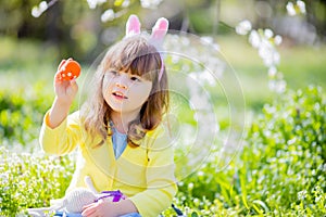 Cute little girl with curly hair wearing bunny ears and summer dress having fun during Easter egg hunt relaxing in the garden