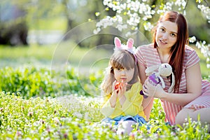 Cute little girl with curly hair wearing bunny ears and summer dress having fun with her young mother, relaxing in the garden